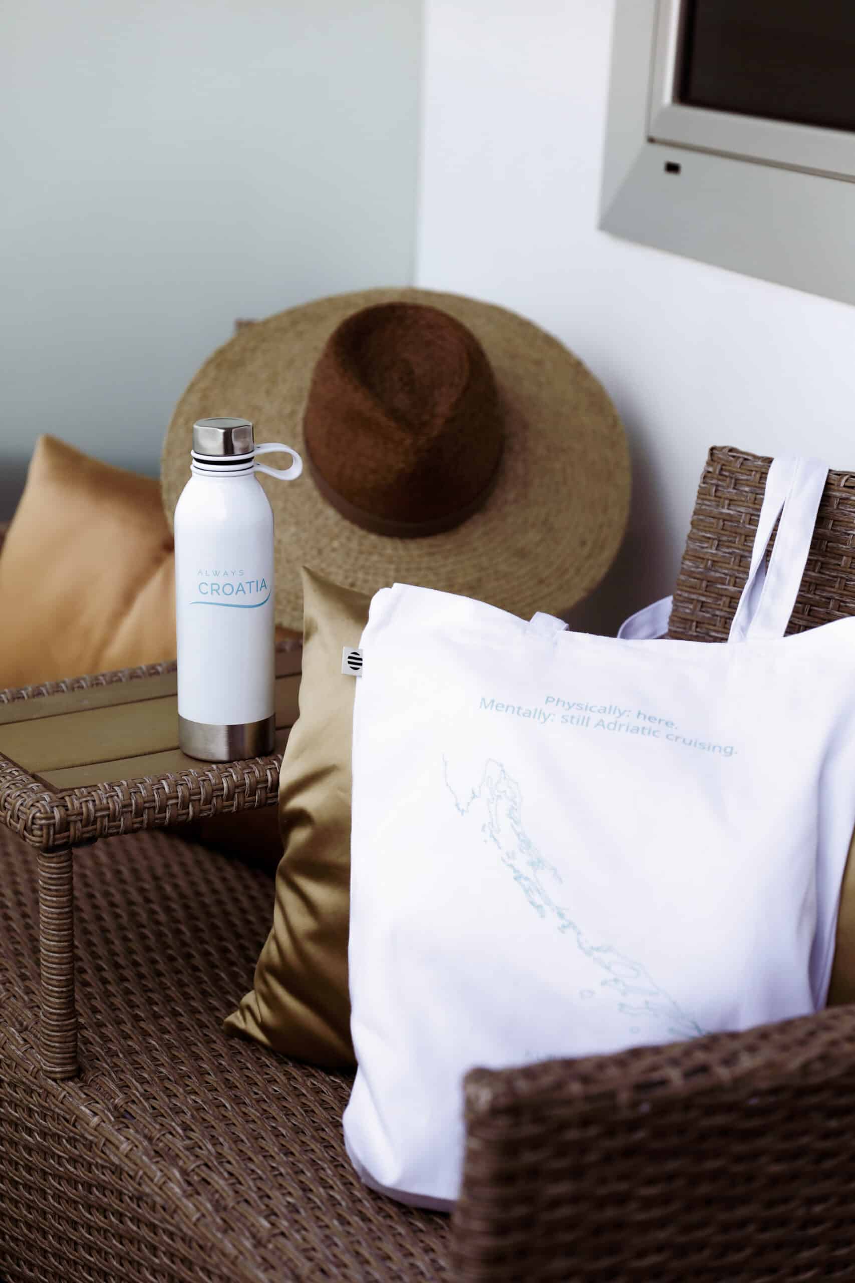 A straw hat, water bottle, and a tote bag with a printed map of Croatia are placed on a wicker chair with cushions, perfect for those embarking on an MS Premier Small Ship Sailing Cruise With Always Croatia.