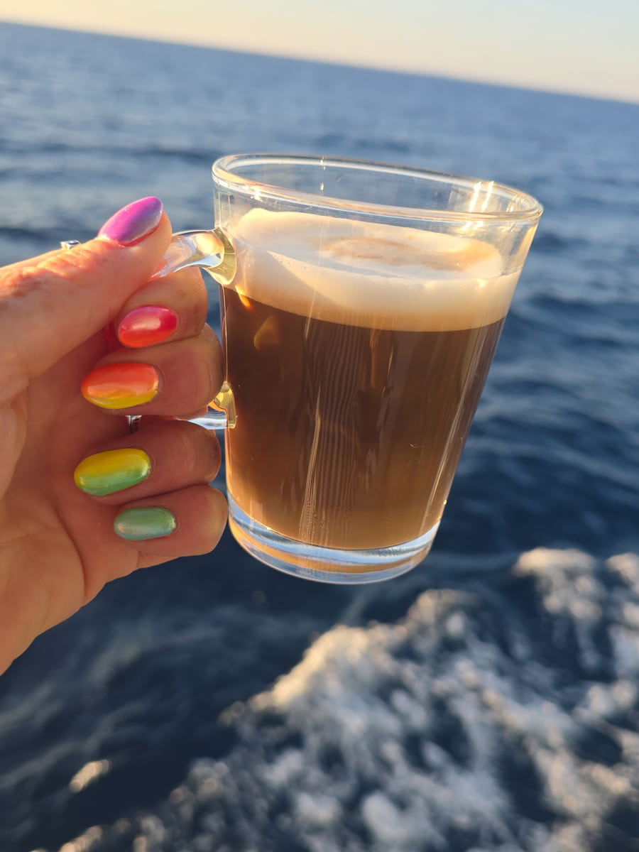 SJ with multicolored nails holds a coffee over the sea and waves from a luxury cruise.