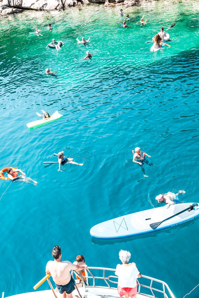 People swimming and paddleboarding in the clear blue waters of the Adriatic Sea, with some using floatation devices. A few individuals are on a boat, watching the activities below.