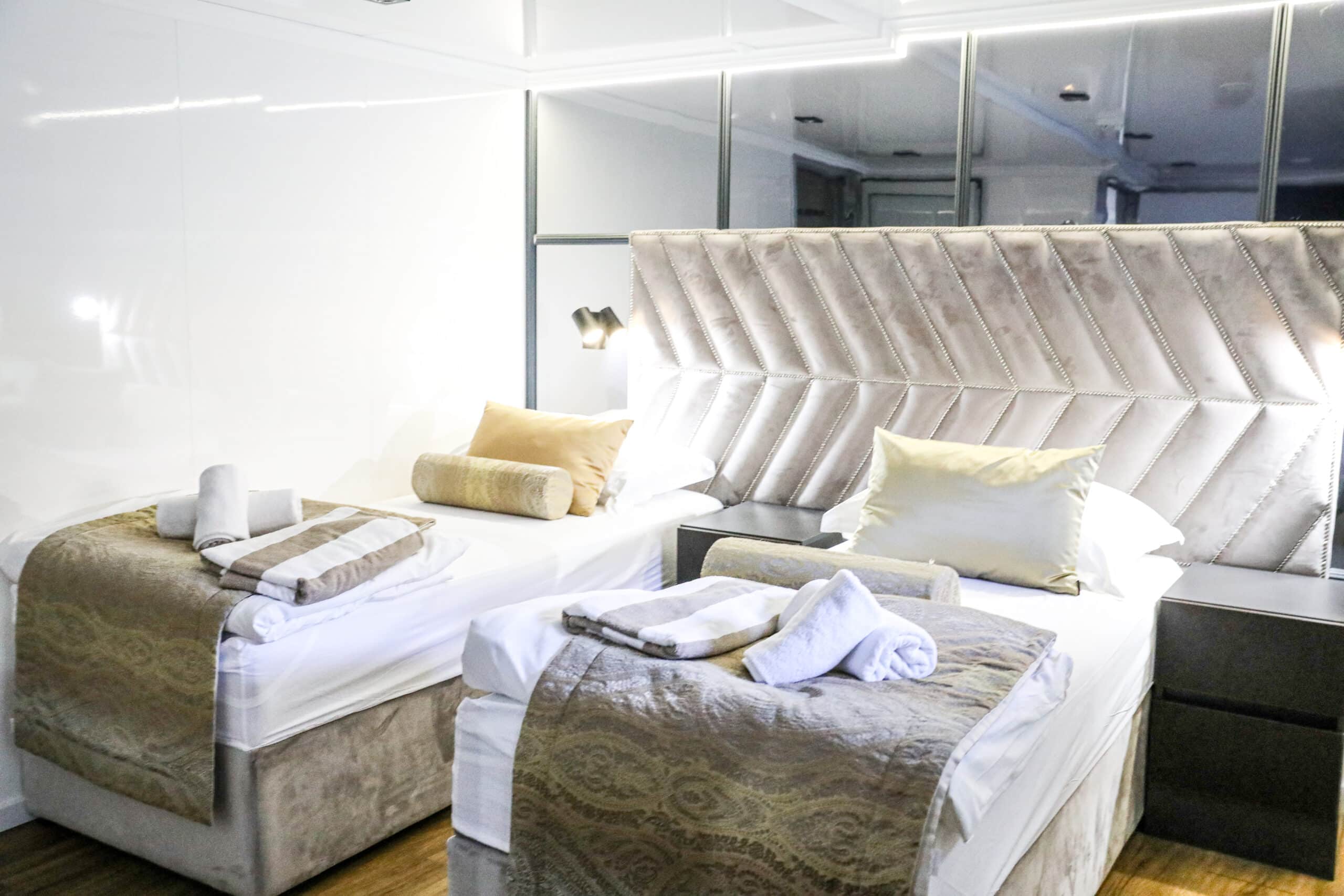 A neatly arranged room with two single beds, each dressed in white linens and beige throws, with rolled towels on top. A padded beige headboard and nightstand with a lamp adorn the space, reminiscent of the luxurious comfort found on an MS Premier Small Ship Sailing Cruise With Always Croatia.